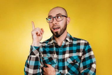 İnsanlar ve duygular. Gözlüklü, kel, sakallı ve mavi gömlekli bir adam yeni bir fikre işaret ediyor. Sarı arka plan. Boşluğu kopyala.