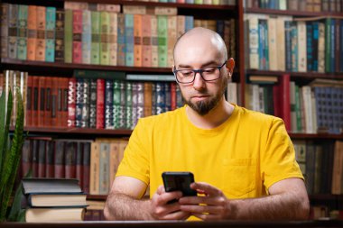 İletişim. Gözlüklü kel bir adam akıllı telefon kullanır. Arka planda bir kütüphane ve kitap rafları var. Sosyal ağlar, uzaklık öğrenme ve karantina kavramı.