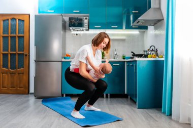 Genç beyaz anne mutfakta çömeliyor, bebeği kollarında tutuyor. Çocuklarla evde spor eğitimi kavramı.
