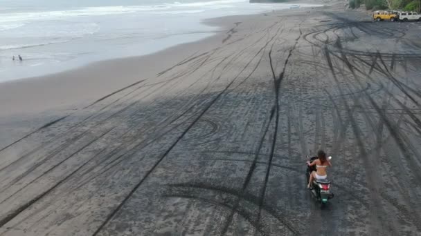 一个穿着泳衣的年轻女子骑摩托车在黑色海滩上的空中照片 背景是黑色海滩和海洋 无人机 后视镜 — 图库视频影像