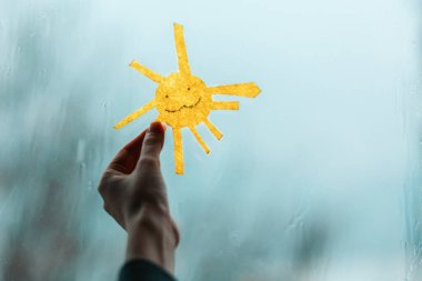 Bir çocuğun eli güneşi tutuyor, keçeden kesilmiş, pencereye karşı. Pencerenin dışında, yağmur ve kötü hava. Yakın plan. Depresyon ve iyi ruh hali kavramı