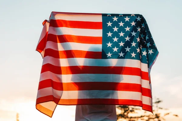 独立日 女人的侧影手里拿着一面美国国旗 高高地高举在头顶之上 后视镜在背景 日落的天空 美国国定假日的概念 — 图库照片