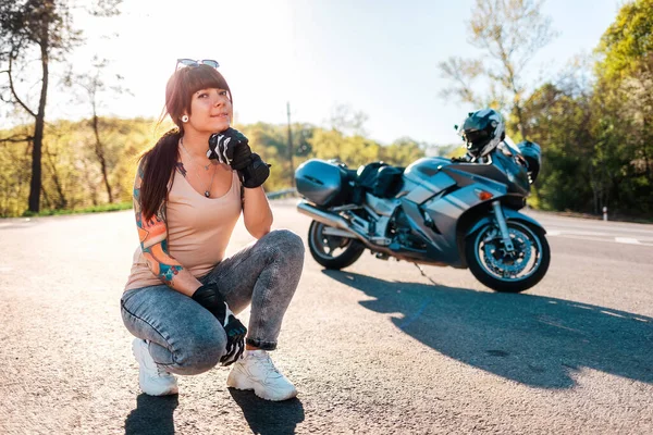 Motorcycle local travel. Smiling young caucasian woman with tattoos on her arm, wearing motorcycle gloves posing sitting. Road and motorbike in the background. World Motorcyclist Day concept.