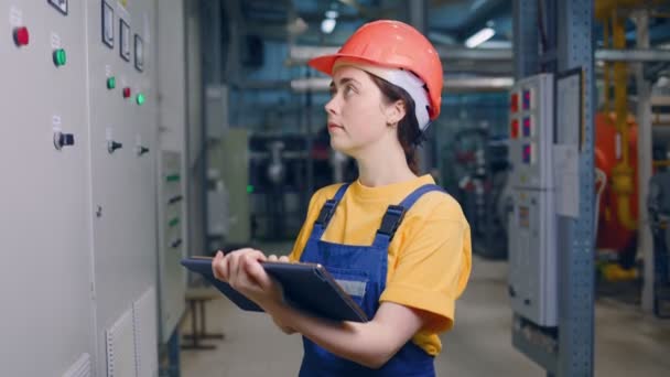 Seorang insinyur perempuan muda dalam helm pelindung, menuliskan pembacaan meteran dalam tablet. Industri dan manufaktur modern — Stok Video