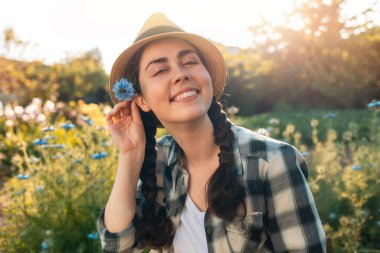 Merhaba Summer. Hasır şapkalı mutlu bir genç kadının portresi kulağının arkasında mavi bir nigella çiçeği tutuyor. Güneş ışığı.