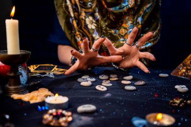 Falcının kadın eli rünleri okur. Tarot kartları, mücevherler ve mumlar masada. Kehanet, astroloji ve geleceği tahmin etme kavramı..