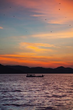 Suda yüzen bir tekne manzarası. Kıyı şeridinin arka planında ve alevli bir günbatımı. Dikey yönelim.