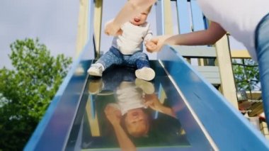 Anne çocuk parkında bir çocukla oynuyor, mutlu kızına çocuk kaydırağını yuvarlamayı öğretiyor. Arka planda oyun alanı.
