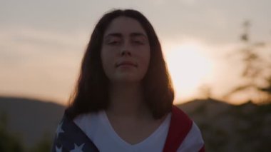 Omuzlarında Amerikan bayrağı olan genç bir kadının portresi, günbatımının arka planında. Ağır çekim. Vatanseverlik kavramı.