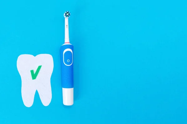 Concept of modern dental care products. Electric toothbrush and a tooth cut out of felt with a green ok icon. Blue background. Flat lay. Copy space.