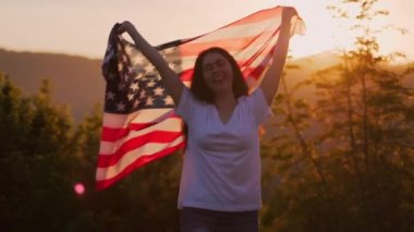 Vatanseverlik. Genç, gülümseyen bir kadın Amerikan bayrağını sallayarak öne çıkıyor. Arka planda günbatımı gökyüzü. Ağır çekim. Amerikan Ulusal Tatil Konsepti.