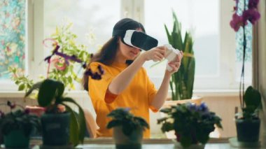  VR gözlüklü duygusal bir kadın ve joystick ile bilgisayar oyunu oynuyor. İçerisi doğal bitkilerle dekore edilmiş. Arka planda bir pencere var. Modern teknoloji kavramı..