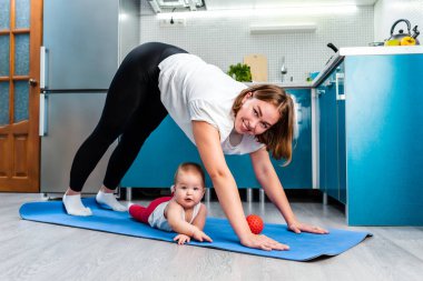 Evde yoga. Beyaz bir anne, bebeği paspasın altındayken Adho Mukha Shvanasana taklidi yapıyor. Çocuklarla spor aktivitesi kavramı.