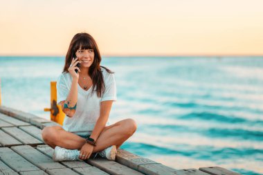 Kolunda dövme olan mutlu bir kadın iskelede oturur ve telefonda konuşur. Arka planda deniz. İletişim ve deniz tatili kavramı.