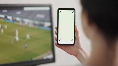 Elinde yeşil ekranlı cep telefonu olan bir kadın. Arka planda futbol maçı yayınlanan bir televizyon ekranı var. Gerçek zamanlı. Spor bahisleri kavramı..