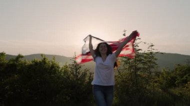 Genç, mutlu bir kadın, Amerikan bayrağını omzunun üzerinden sarkıtarak atlıyor. Arka planda günbatımı gökyüzü. Ağır çekim. Amerikan Ulusal Tatil Konsepti.