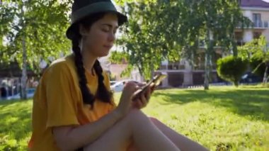 Akıllı telefon kullanan ve kameraya bakan gülümseyen genç bir kadının portresi. Arka planda güneşli bir ışık. Gerçek zamanlı. Çevrimiçi iletişim ve internet bağımlılığı kavramı.