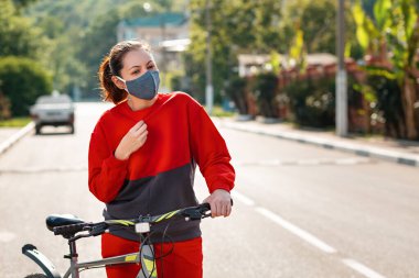 Virüsten koruma. Koruyucu tıbbi maske ve spor giysili bir kadının portresi. Bisiklet tutuyor, ateşi ve nefes darlığı çekiyor. Açık havada. Virüs salgını kavramı.