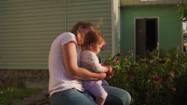 Summertime. A happy young mother holds her toddler at arms and looks together on a flower cut from flower bed in the backyard. Zoom in.