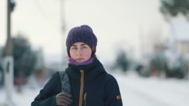 A young woman in a jacket and hat is standing on the street. In the background, a snow-covered street. Winter. Slow motion.
