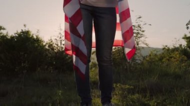 Vatanseverlik. Omzuna Amerikan bayrağı sarılmış genç bir kadın günbatımı gökyüzünün arka planına doğru kaldırıyor ve sallıyor. Flok yelkeni. Ağır çekim. Bağımsızlık Günü kavramı.