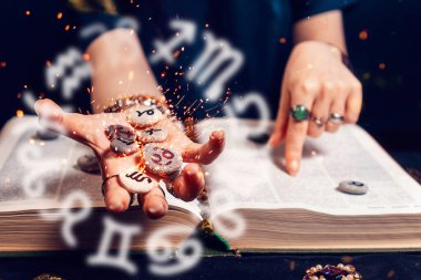 Astroloji ve yıldız falı. Falcının eli, parlak zodyak taşlarını açık kitabın üzerinde avucunun içinde tutuyor. Kehanet ve sihir kavramı.