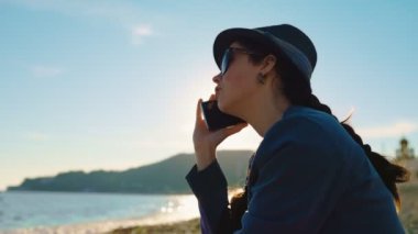 Genç ve güzel bir kadın okyanusun sahilinde oturuyor ve telefonda konuşuyor. Yan görüş. Cep telefonuyla iletişim konsepti.
