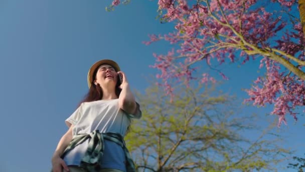 지푸라기 모자를 여자가 아래서서 전화로 이야기를 아래는 꽃피는 배경에 — 비디오