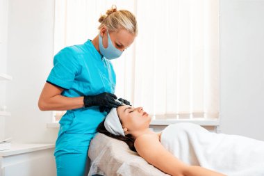 Kadın kozmetoloji salonunda alnına iğne yaptırıyor. Lateks eldivenli ve koruyucu maskeli kozmetolojist hiyalüronik asit şırıngası yapıyor. Estetik ameliyat ve yüz gerdirme kavramı
