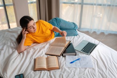 Eğitim. Genç bir kadın yatağında uzanmış kitap okuyor. Laptop ve belgeler yan yana duruyor. Üst Manzara. Çevrimiçi kurslar kavramı.