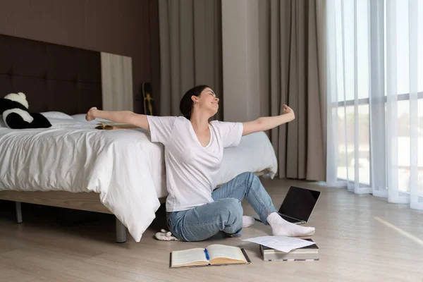 A young woman sits on the floor of a room and stretches after a long training session. The books are on the bed and on the floor. The concept of online education.