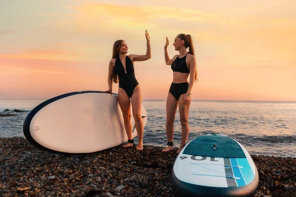 2人の幸せな女性がボードを持ち 5人の手でビーチに立っています 背景には 海と夕日 夏の極端なレクリエーションとサーフィン — ストック写真