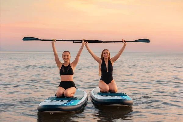 两个快乐的女人坐在一个冲浪板上 举起桨 在背景中 海洋和日落 夏季极限运动 — 图库照片
