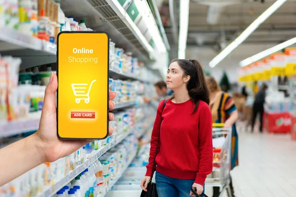 Concept of online shopping. A young pretty woman looking at the display cases with dairy products in the supermarket refrigerator. The hand holds the mobile phone on the left.