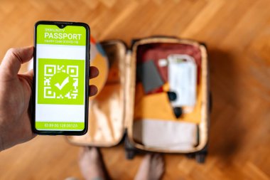 Yeni normal kavramı. Erkek eli, bağışıklık sistemi dijital pasaportlu bir akıllı telefon tutuyor. Bulanık arka planda elbiseli ve koruyucu maskeli bir bavul..