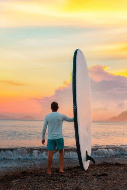 Sup tahtası tutan ve sahilde poz veren formda bir adam. Arka planda, okyanus ve günbatımı. Dikey. Uzayı kopyala Yaz sörfü ve aşırı etkinlik.
