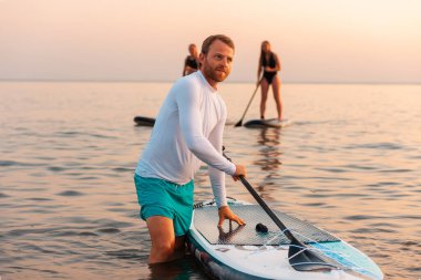 Yemek tahtasıyla yüzen adam. Arka planda okyanusta sörf yapan iki kadın. Yaz sporu ve eğlencesi.