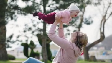 Genç mutlu bir anne çimlerin üzerinde oturuyor, bebeğiyle oynuyor. Gerçek zamanlı.