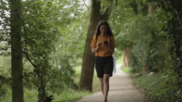 若い女性が公園内の道を歩き スマートフォンでタイプします 本物の景色だ オンライン中毒の概念 — ストック動画