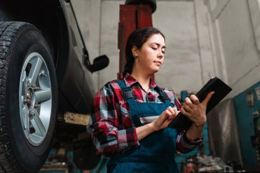 Üniformalı, elinde tablet olan beyaz bir kadın tamircinin portresi. Arabanın tekerleği sağda. Alt görünüm.