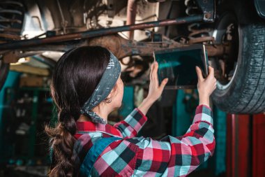 Araba tanısı koymak için tablet kullanan, asansörde arabanın altında duran üniformalı genç bir kadın tamircinin portresi. Arka görünüm.