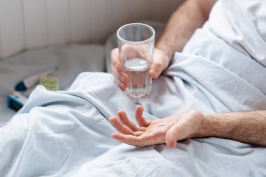 Bir adam elinde bir bardak suyla yatakta uzanıyor ve elinde bir hap tutuyor. Üst Manzara. Tedavi kavramı.
