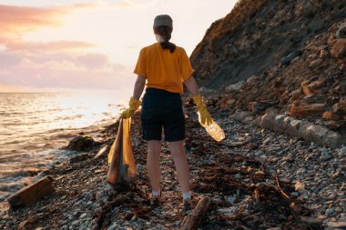 Plastik şişe tutan ve kumsalda poz veren kadın gün batımını seyrediyor. Arkadan bak. Dünya Günü ve çevresel koruma kavramı.
