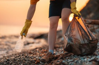 Dünya günü. Okyanus kıyısındaki çöpleri temizliyorum. Plajdan plastik bir şişe alan polyethielene çantalı bir gönüllü. Yakın plan. Arka planda gün batımı. Ekolojinin korunumu kavramı.