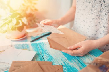 Bir kadın kartpostal okuyor. Harfi harfine yakın çekilen eller. Arkadaki mavi ahşap masa. Işık. Geçiş sonrası ve tatil tebrikleri..
