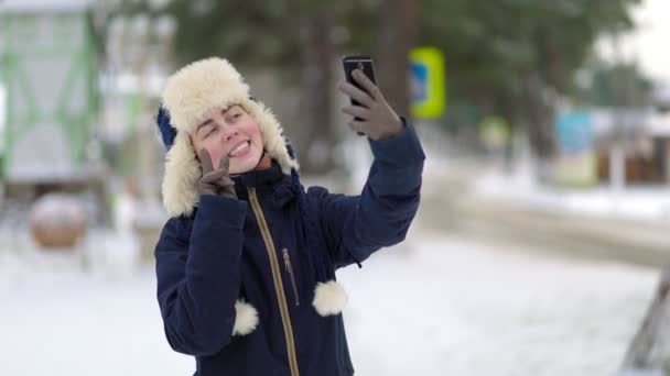 スマートフォンで自撮りをする耳フラップとジャケットを持った帽子を被った若い笑顔の女性の肖像 背景に雪に覆われた通り — ストック動画