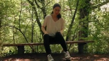 Genç bir kadın bankta oturuyor, sivrisinek ısırıklarından kendini tırmalıyor. Arka planda yaz ormanı var. Gerçek zamanlı. Çekim.