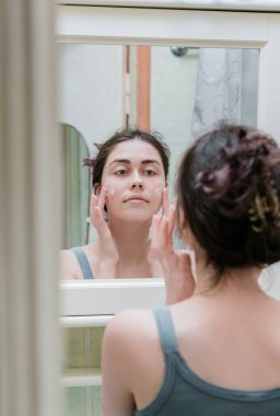 Güzel kız aynanın önünde yüzüne dokunuyor ve cildini kremle nemlendiriyor. Mükemmel saf ten..