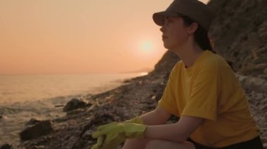 Dünya Günü. Plastik eldivenli bir kadın gönüllü kıyıda oturuyor, sahili temizledikten sonra dinleniyor. Kıyı bölgelerini çöpten temizleme kavramı.