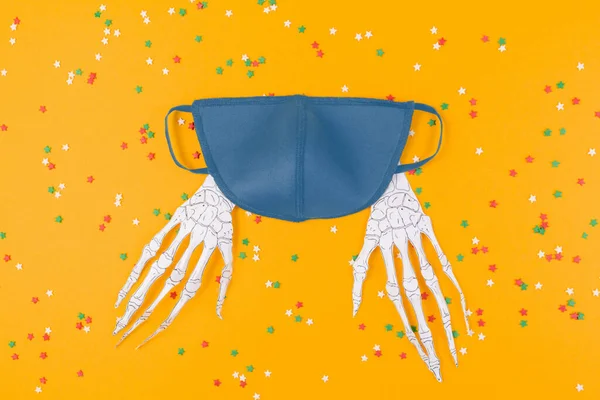 Reusable blue virus protection mask and paper skeleton hands. Orange background with decorative stars. Flat lay. The concept of protection from the virus during Halloween.